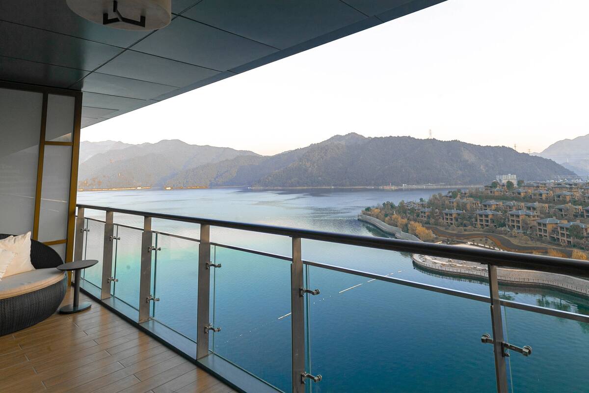 A balcony with a view of a river and mountains, a wicker sofa, and a small table.