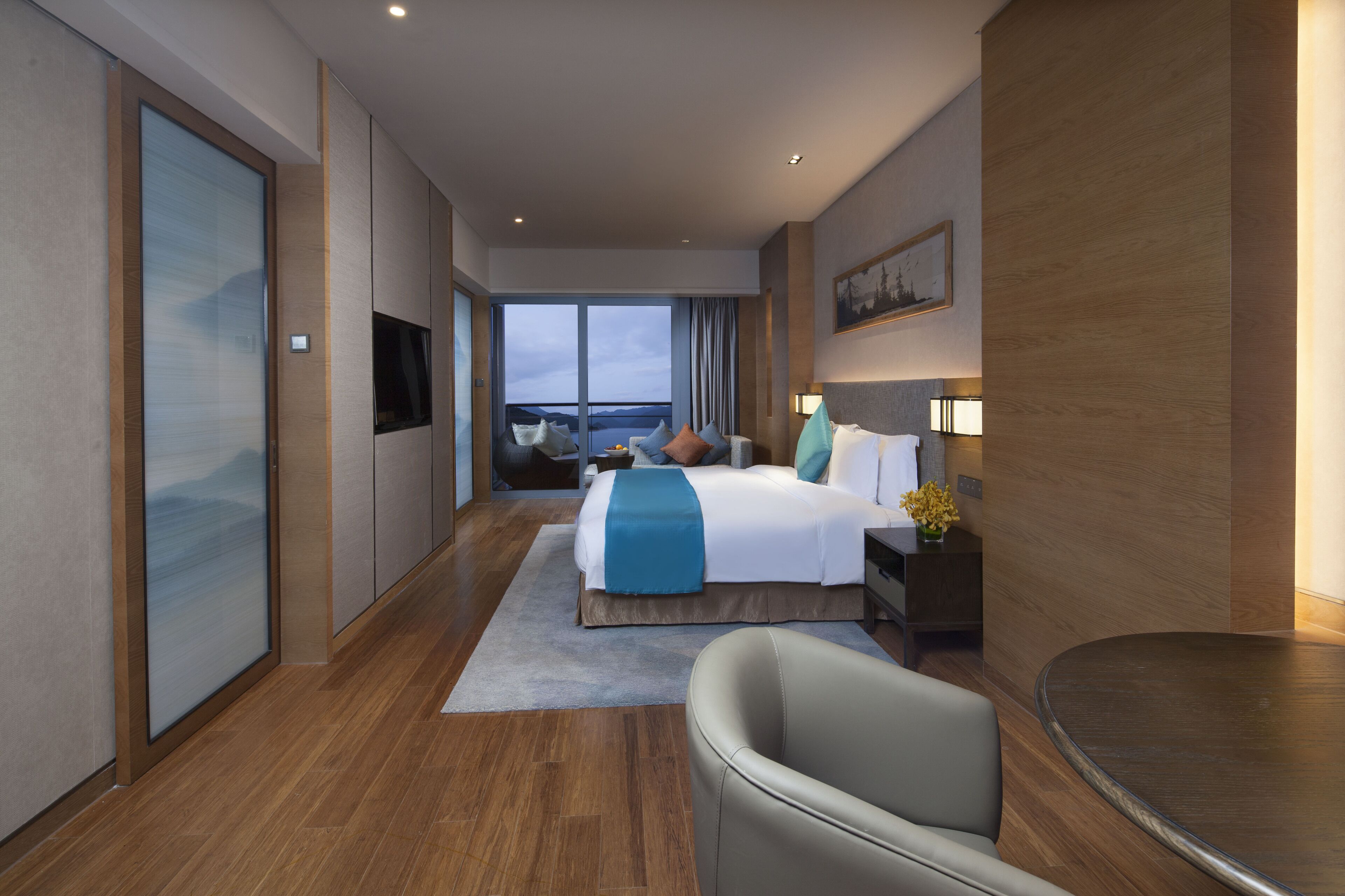 A modern hotel room with a large bed, a seating area, a TV, and a view of the outdoors.