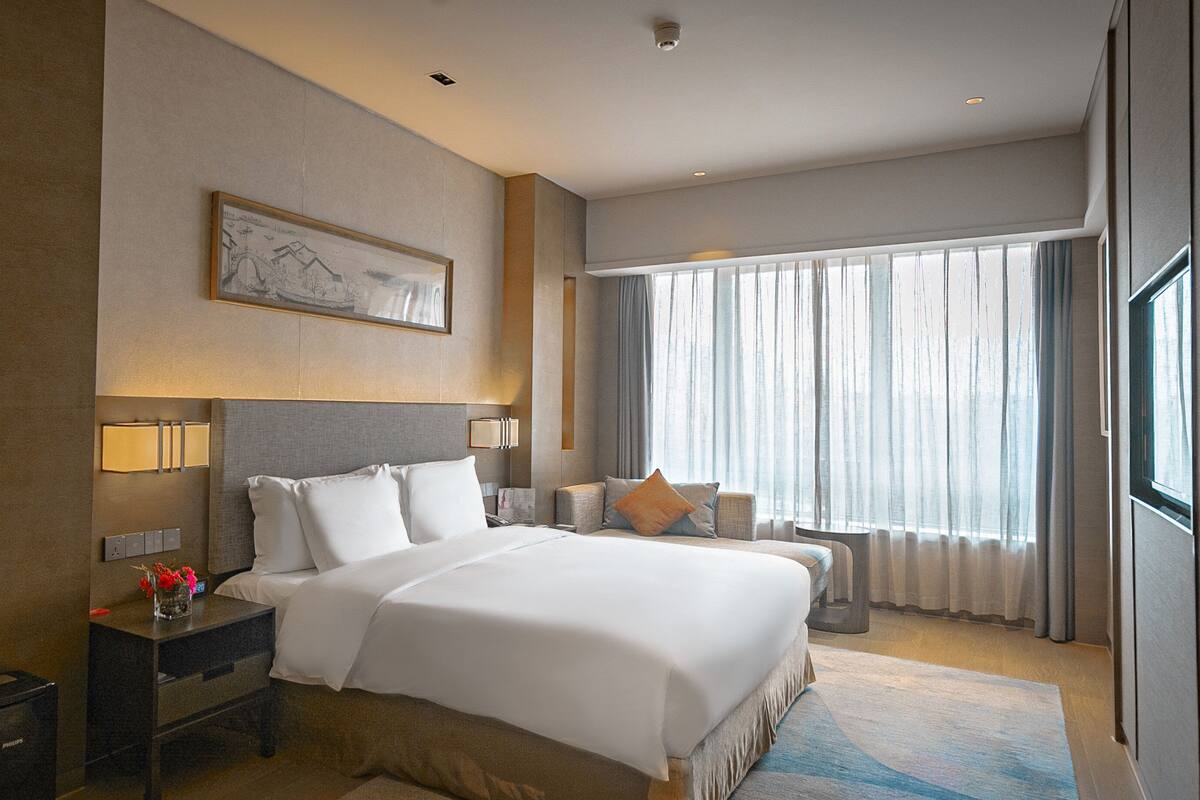 A hotel room with a large bed, bedside tables, a nightstand, a framed picture on the wall, and a window with curtains.