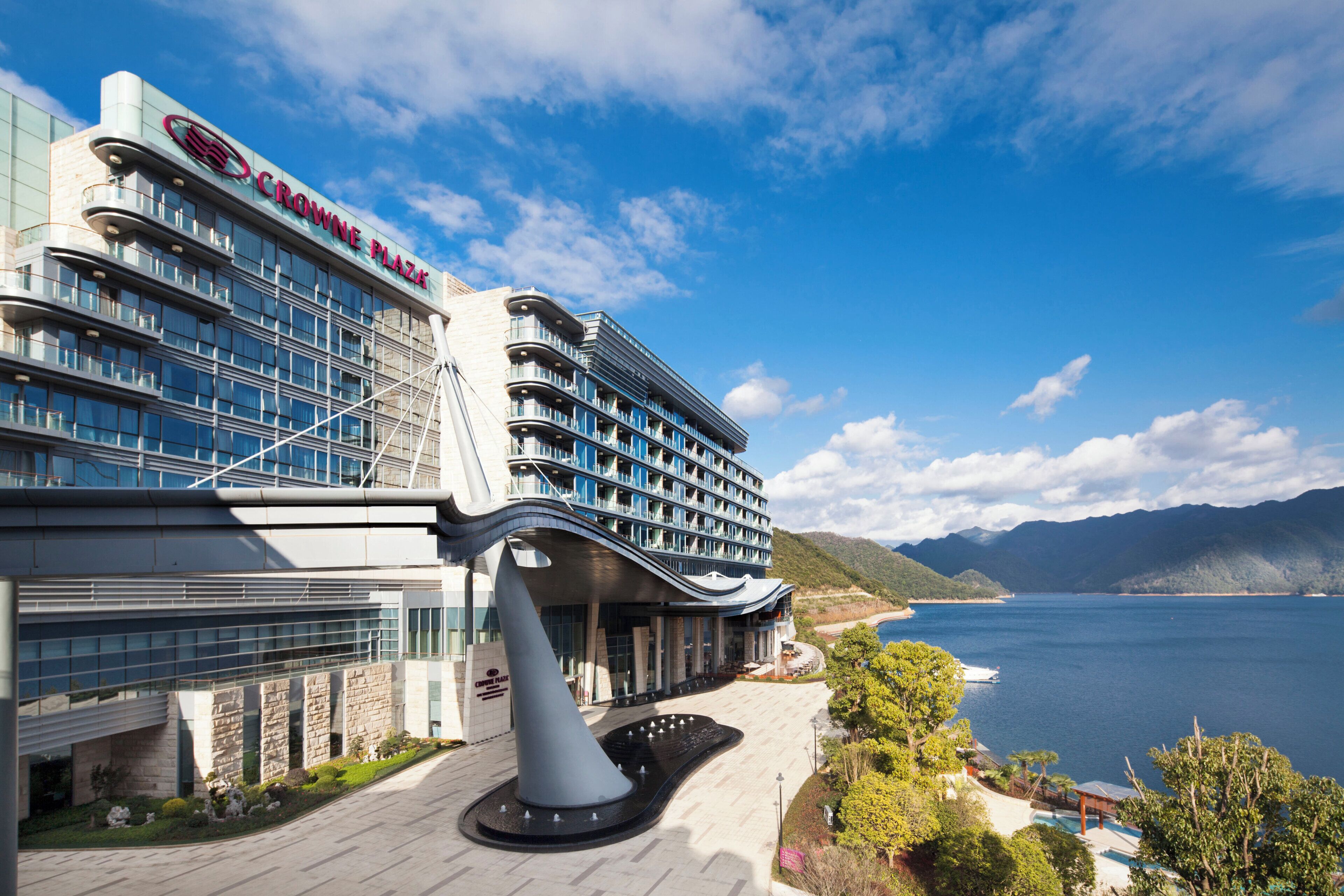 Un hotel moderno con un llamativo letrero 'CROWNE PLAZA', vistas al agua y una zona ajardinada con una fuente.