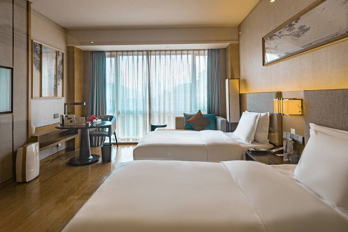 A hotel room with two beds, a desk, a TV, and a large window with curtains.