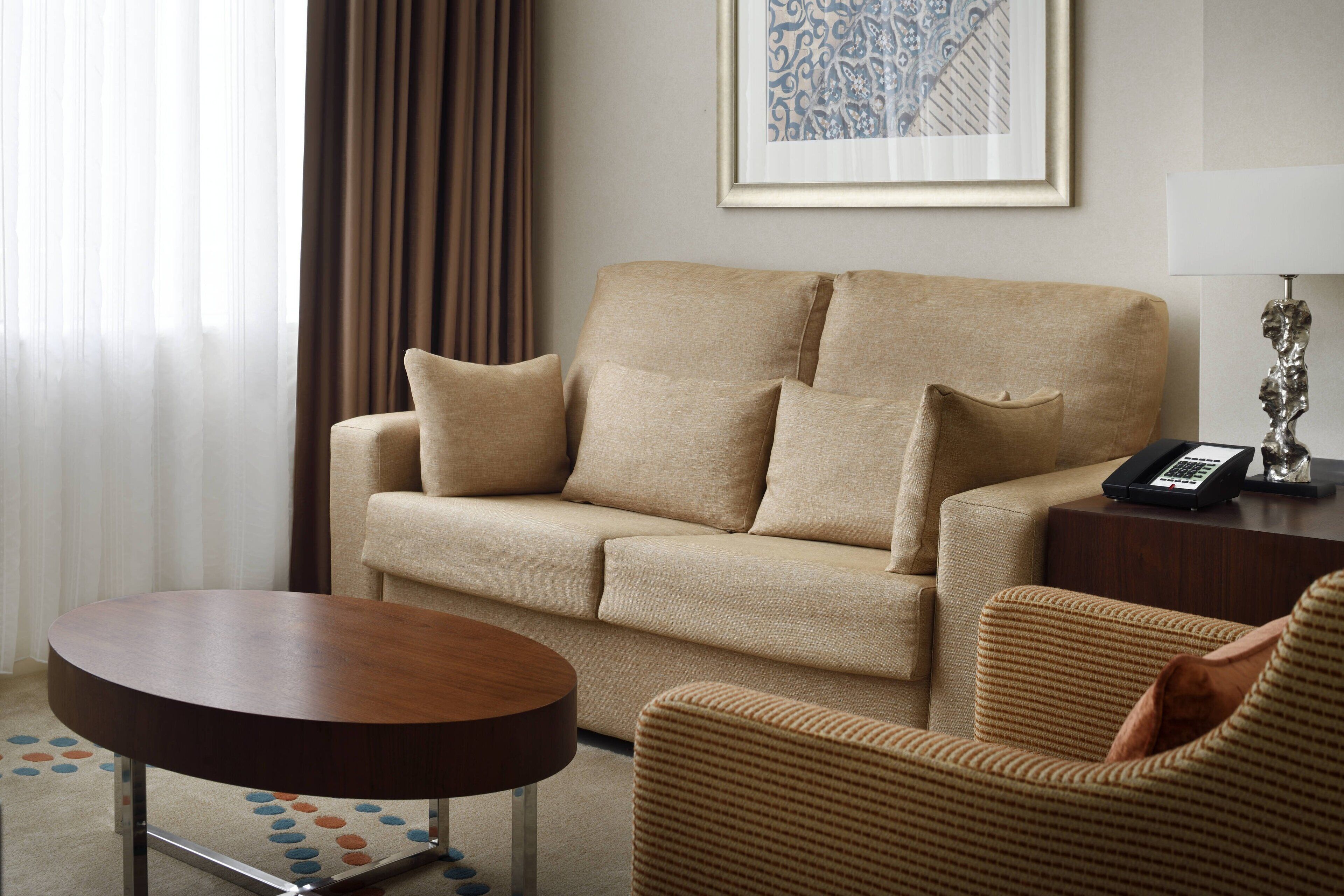 A hotel room with a beige sofa, a round wooden coffee table, a wicker armchair, and a framed artwork on the wall.