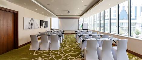 A conference room with a green carpet, white chairs, tables, a projector screen, and a whiteboard.