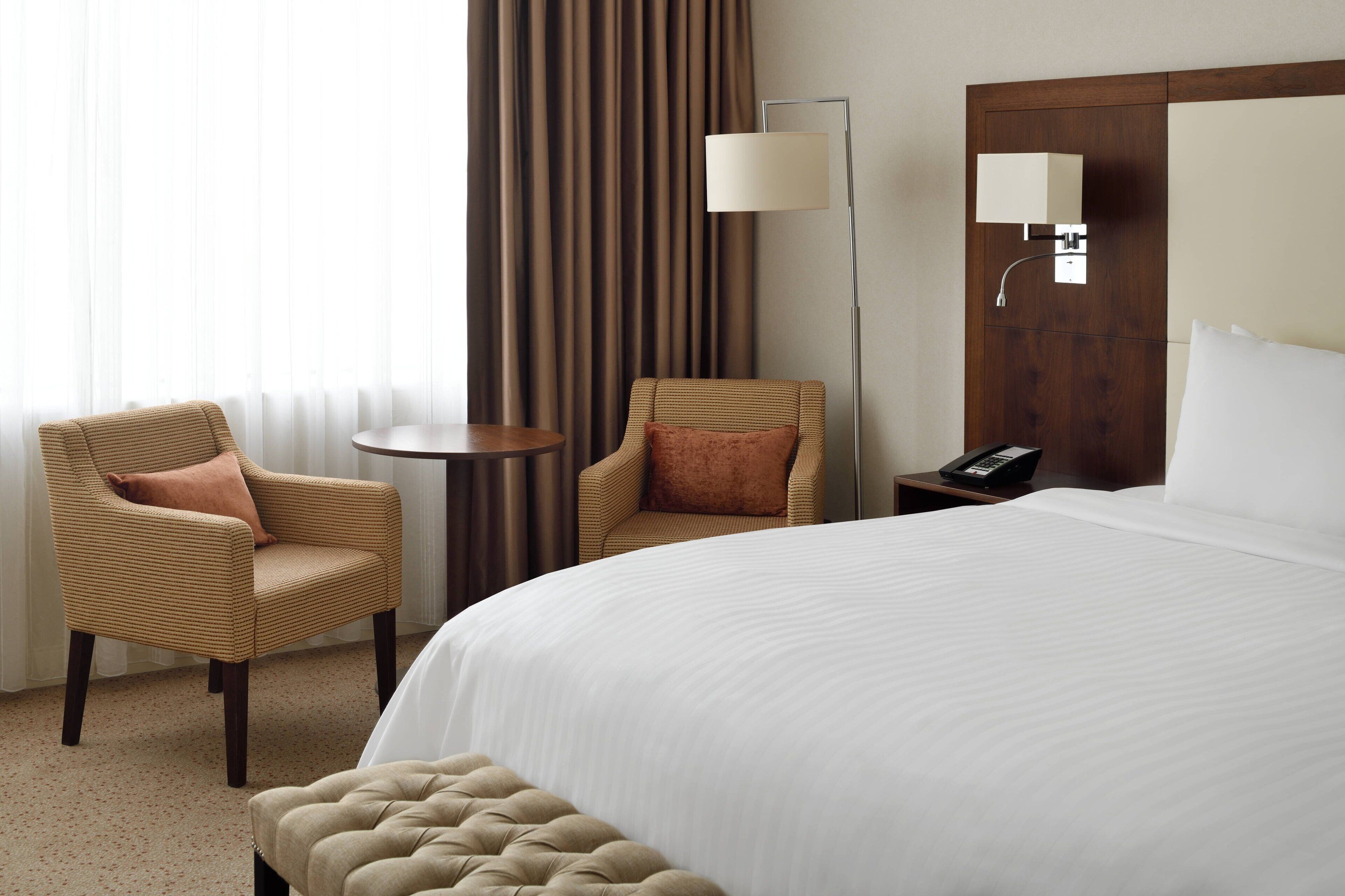 Una habitación de hotel con una cama grande, dos sillones, una mesita redonda pequeña, una lámpara y un teléfono sobre una mesita de noche.