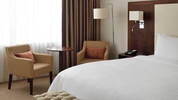Una habitación de hotel con una cama grande, dos sillones, una mesita redonda pequeña, una lámpara y un teléfono sobre una mesita de noche.