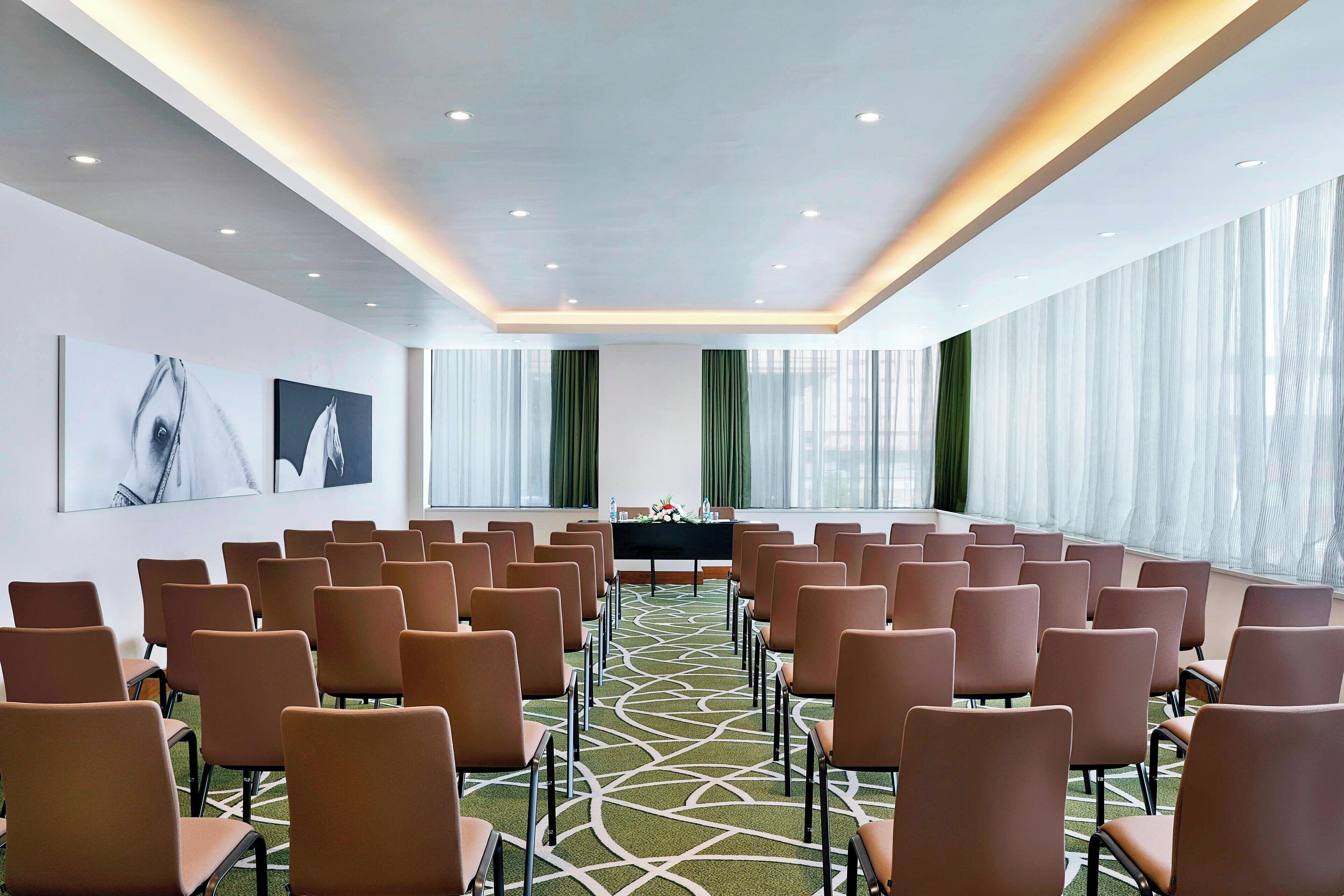 A conference room with rows of chairs, a table with floral centerpieces, and large windows with sheer curtains.
