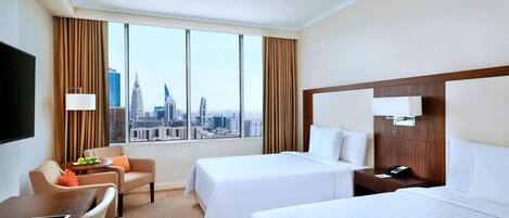 A hotel room with two beds, a desk with a tablet, a chair, a nightstand with a phone, and a large window with a city view.
