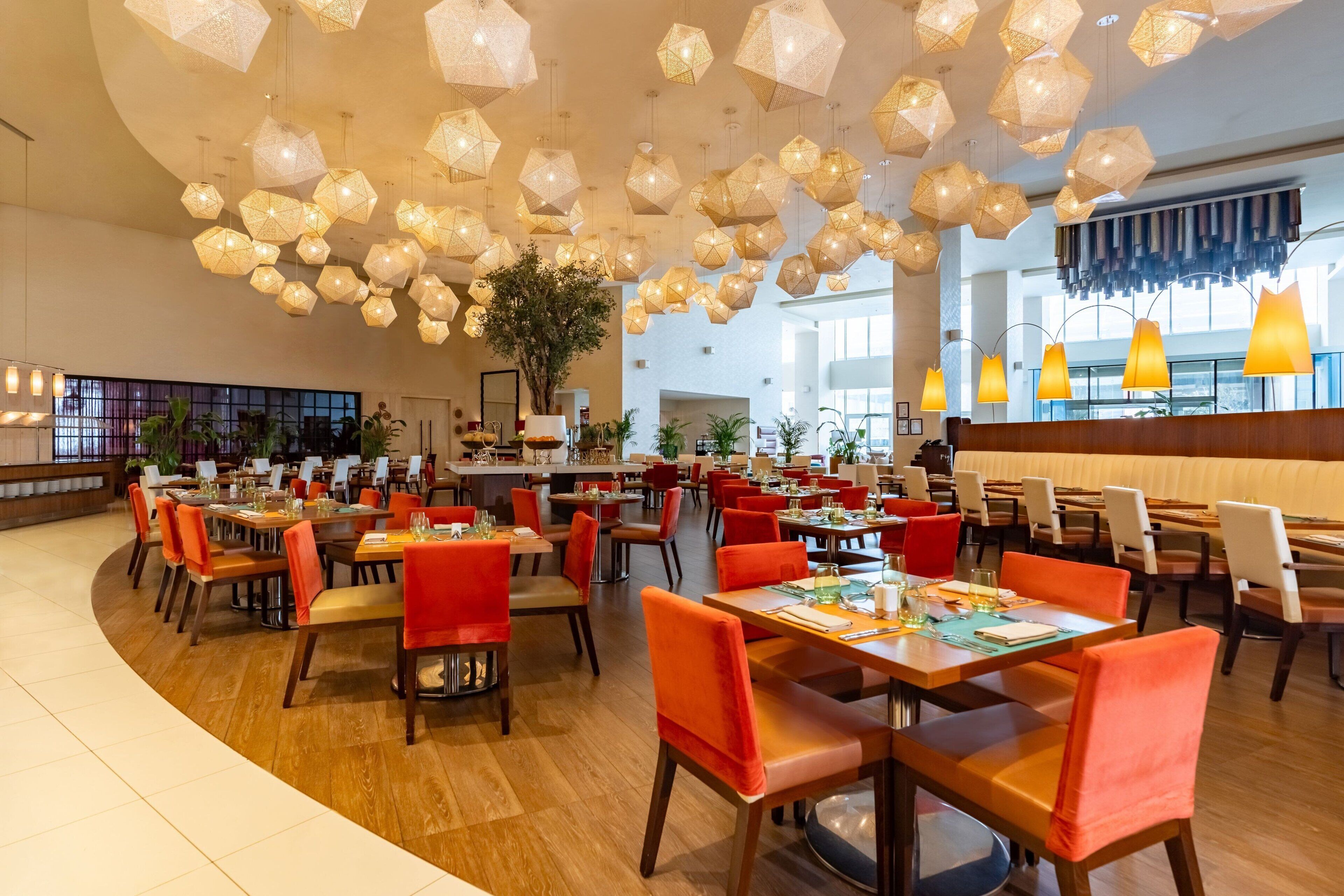 A spacious restaurant with multiple dining tables and chairs, a variety of hanging light fixtures, and large windows allowing natural light.
