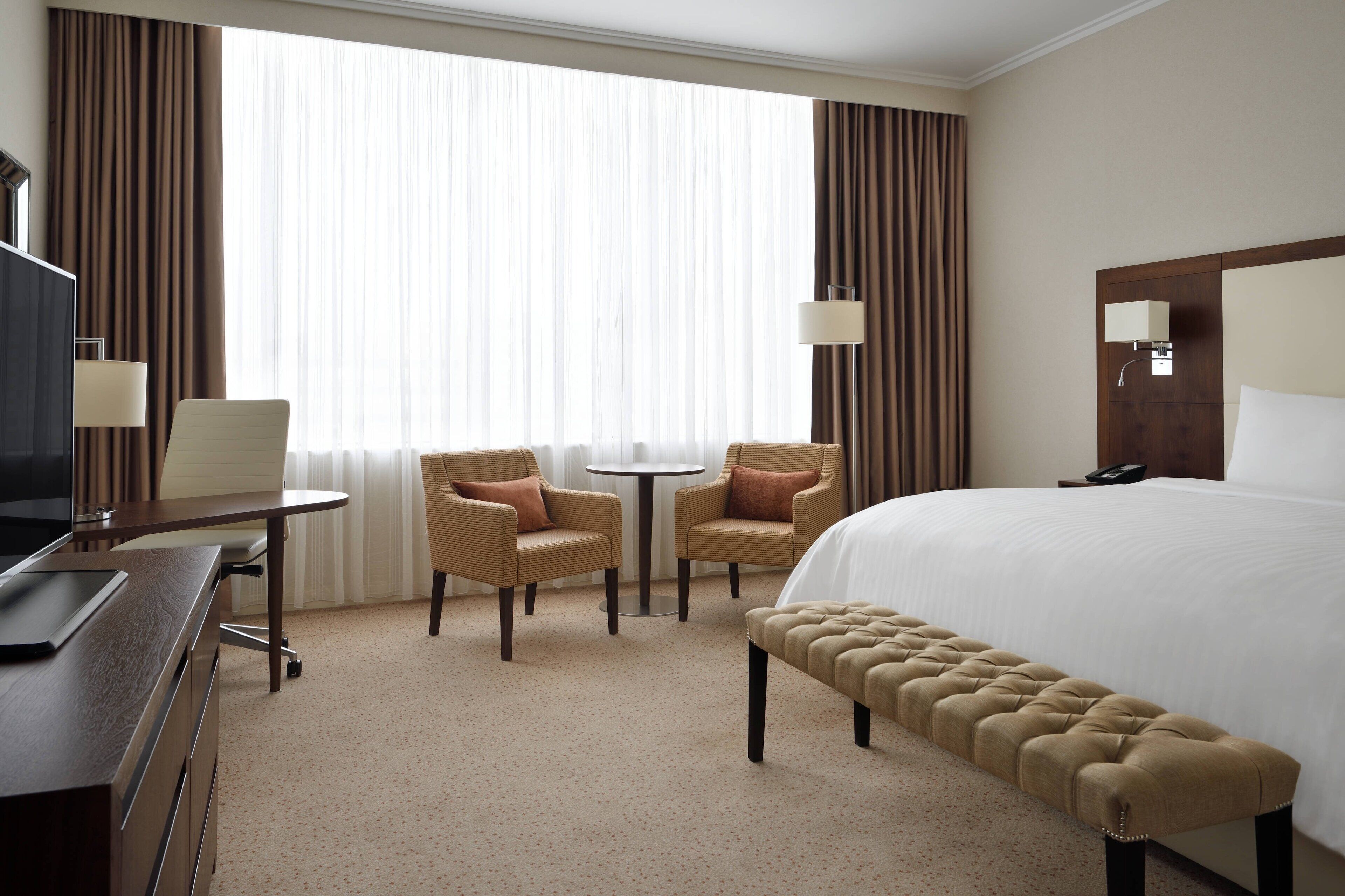 A hotel room with a large bed, two armchairs, a small round table, a desk with a computer, and a TV.