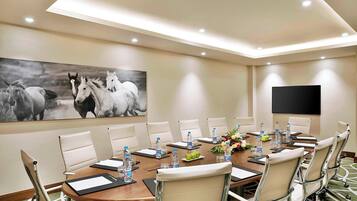 Un salón de conferencias con una larga mesa, sillas blancas, botellas de agua y un cuadro grande de caballos en la pared.