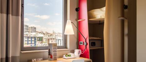 Habitación de hotel compacta con escritorio, silla y cama con cabecera roja. Una ventana ofrece vistas a la ciudad, y hay una lámpara sobre el escritorio.