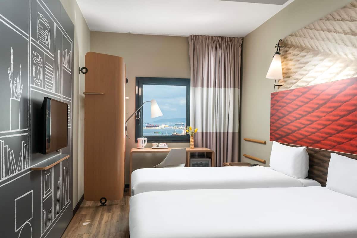 Habitación de hotel con cama, escritorio con lámpara, televisor montado en la pared y ventana con vista al mar.
