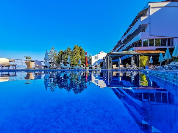 Una piscina azul clara con sillas de descanso y sombrillas, rodeada de edificios modernos y árboles.