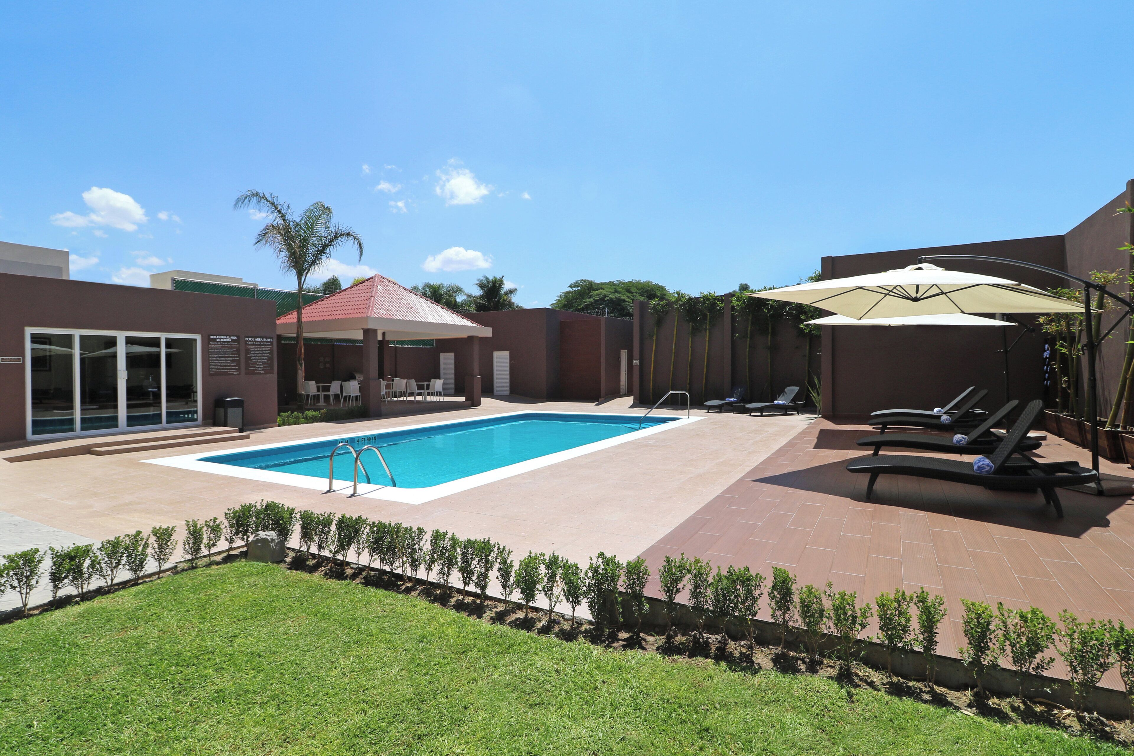 A swimming pool with lounge chairs and umbrellas, surrounded by a brick patio and a grassy area.