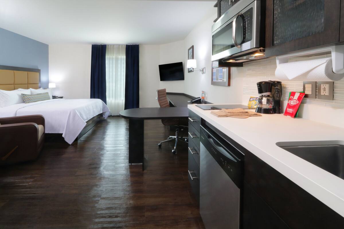 Een kleine hotelkamer met een bed, een bureau, een stoel, een magnetron, een koffiezetapparaat en een vaatwasser.