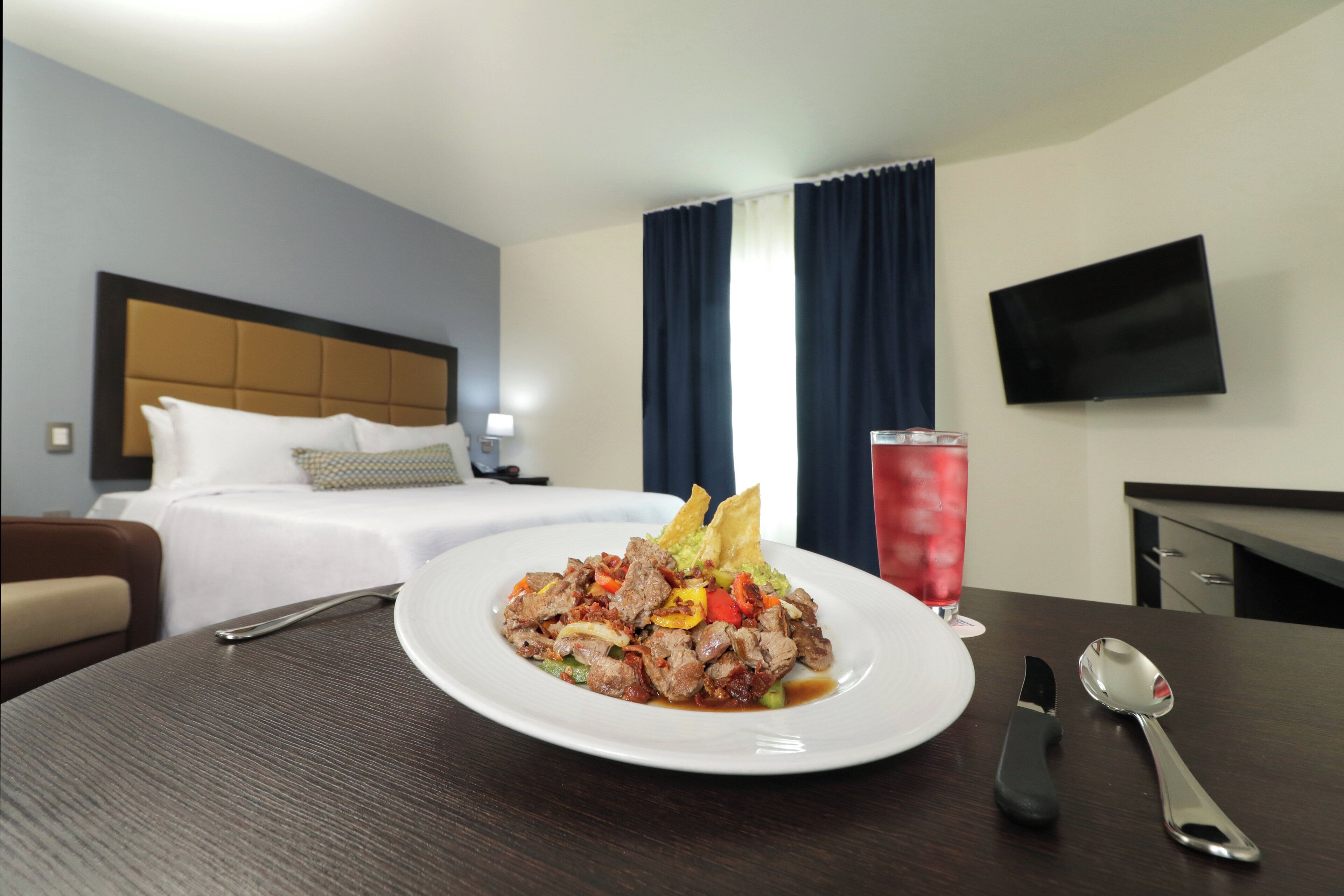 A hotel room with a dining table set for a meal, a bed with a headboard, a TV mounted on the wall, and a window with curtains.