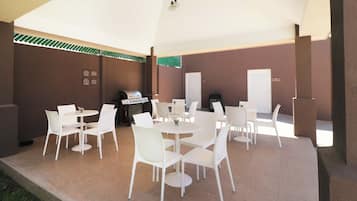 Eine Terrasse im Freien mit weißen Tischen und Stühlen, einem Grill und einem überdachten Bereich.