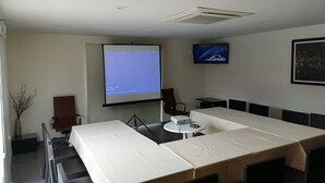 Un salón de conferencias con una gran mesa, sillas, una pantalla de proyector, un televisor y un cuadro en la pared.