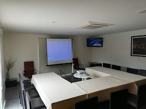 Un salón de conferencias con una gran mesa, sillas, una pantalla de proyector, un televisor y un cuadro en la pared.