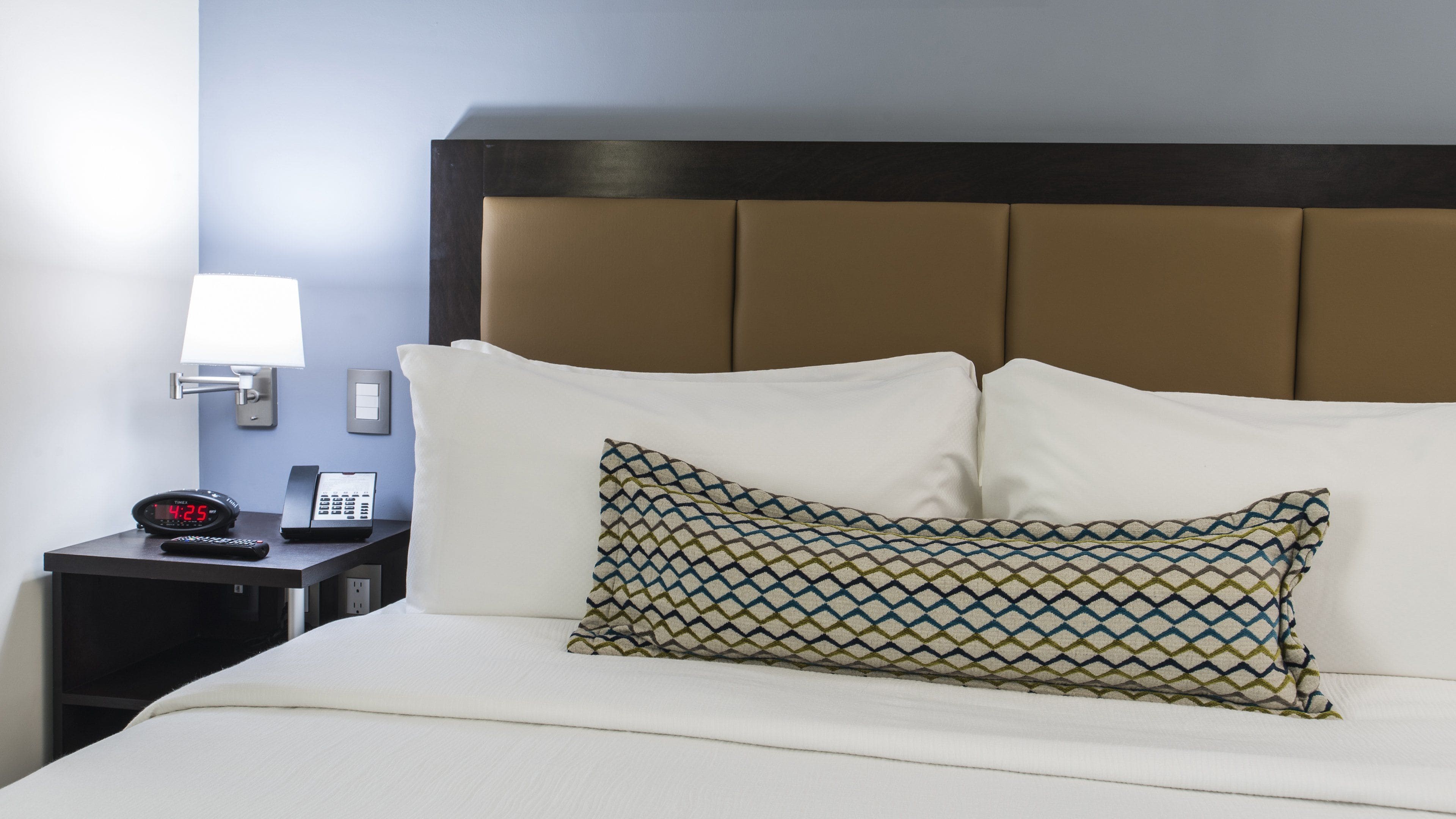 Uma cama bem arrumada com uma almofada estampada, uma mesa de cabeceira com telefone e relógio digital, e uma luminária fixada na parede.