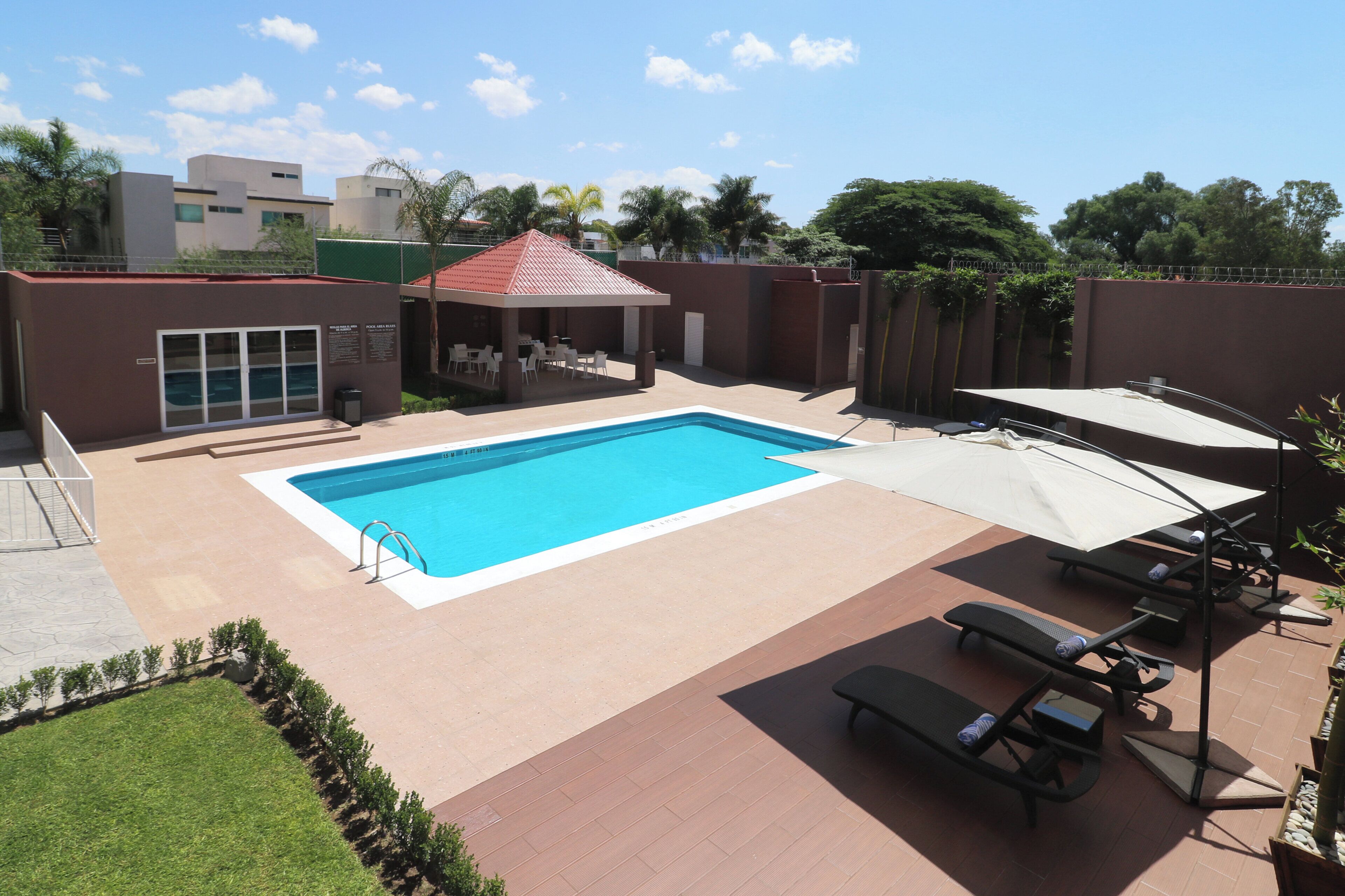 A rectangular swimming pool with a tiled deck surrounding it, a covered seating area with tables and chairs, and a fenced perimeter.