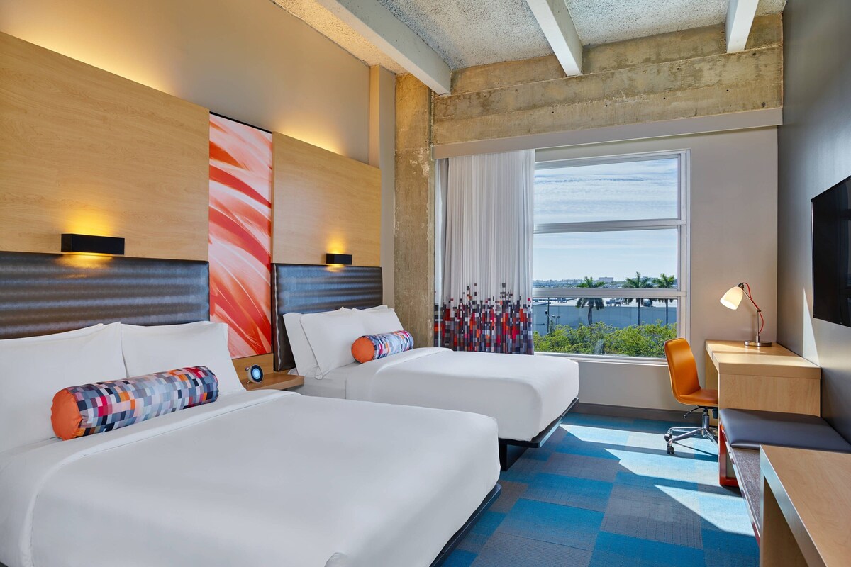 Een hotelkamer met twee bedden, een bureau, een stoel en een groot raam met uitzicht.
