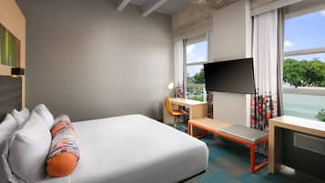 Une chambre d’hôtel équipée d’un lit, d’un bureau, d’une chaise, d’une télévision et d’une fenêtre donnant sur la verdure.