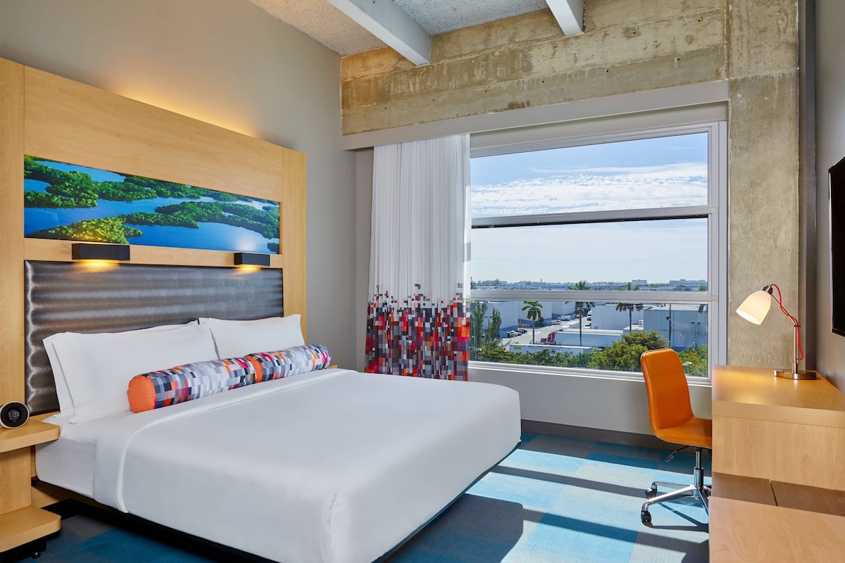 A hotel room with a large bed, a desk, a chair, a window with a city view, and a painting on the wall.
