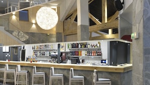Un espace bar moderne avec un plafond haut, des poutres en bois et une mezzanine. On y trouve des tabourets de bar et des tables avec des coussins, ainsi qu’une variété de bouteilles et de verres exposés sur des étagères.