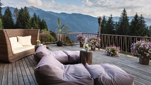 A deck with a wicker sofa and cushions, surrounded by potted flowers and a railing overlooking a mountainous landscape. - Frieden (Pill)