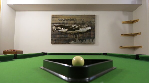 Une table de billard avec une bille blanche au centre, une œuvre d’art représentant un paysage urbain accrochée au mur et des étagères en bois sur le côté droit.