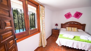 A bedroom with a wooden door, a window with a curtain, a bedside table with a lamp, a bed with a green bedspread, and two wall-mounted rose decorations. - Conjunto Rural Las Rosas (Frigiliana)