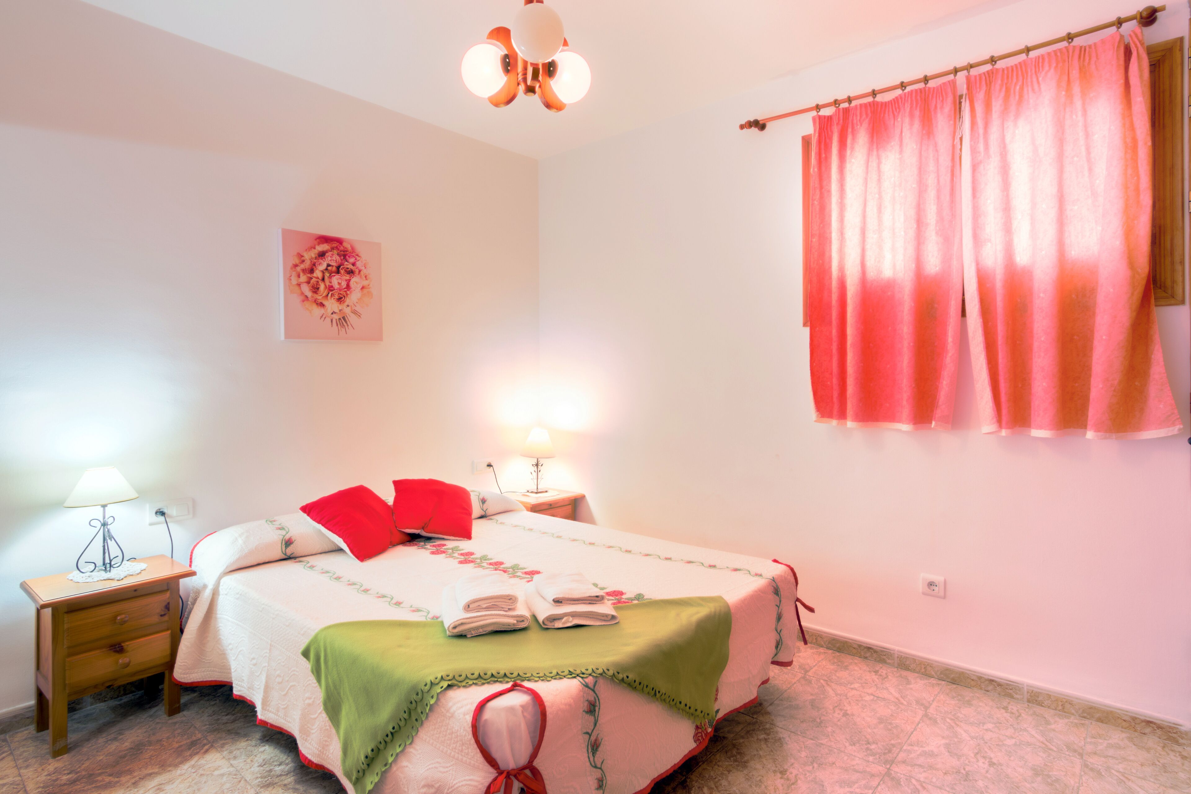 A neatly made bed with a green and white bedspread, two bedside lamps, a floral artwork on the wall, and a pink curtain.
