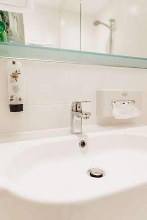 A bathroom with a white sink, a glass shelf above it, a towel dispenser, and a mirror.