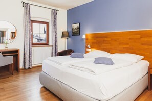 A double bed with white linens and two pillows, a wooden headboard, a desk with a chair, a lamp, and a window with curtains.