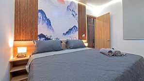 A bedroom with a bed, pillows, a nightstand, a lamp, a wall with a large painting, and a door. - Hotel NG (Cartagena)