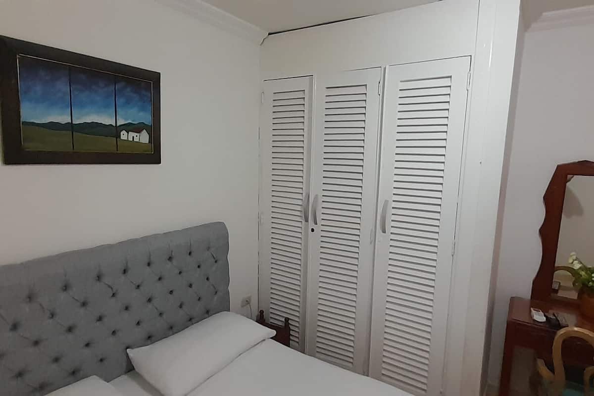 A bedroom with a grey tufted headboard, a white bed, a painting of a landscape, white louvered closets, and a wooden dresser with a mirror.