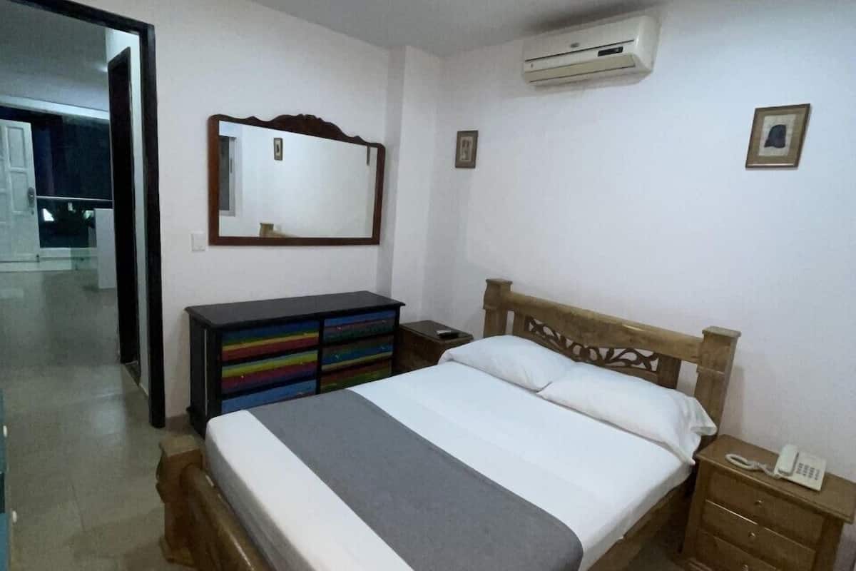 A bedroom with a wooden bed, a nightstand with a phone, a dresser, a mirror, and an air conditioning unit.