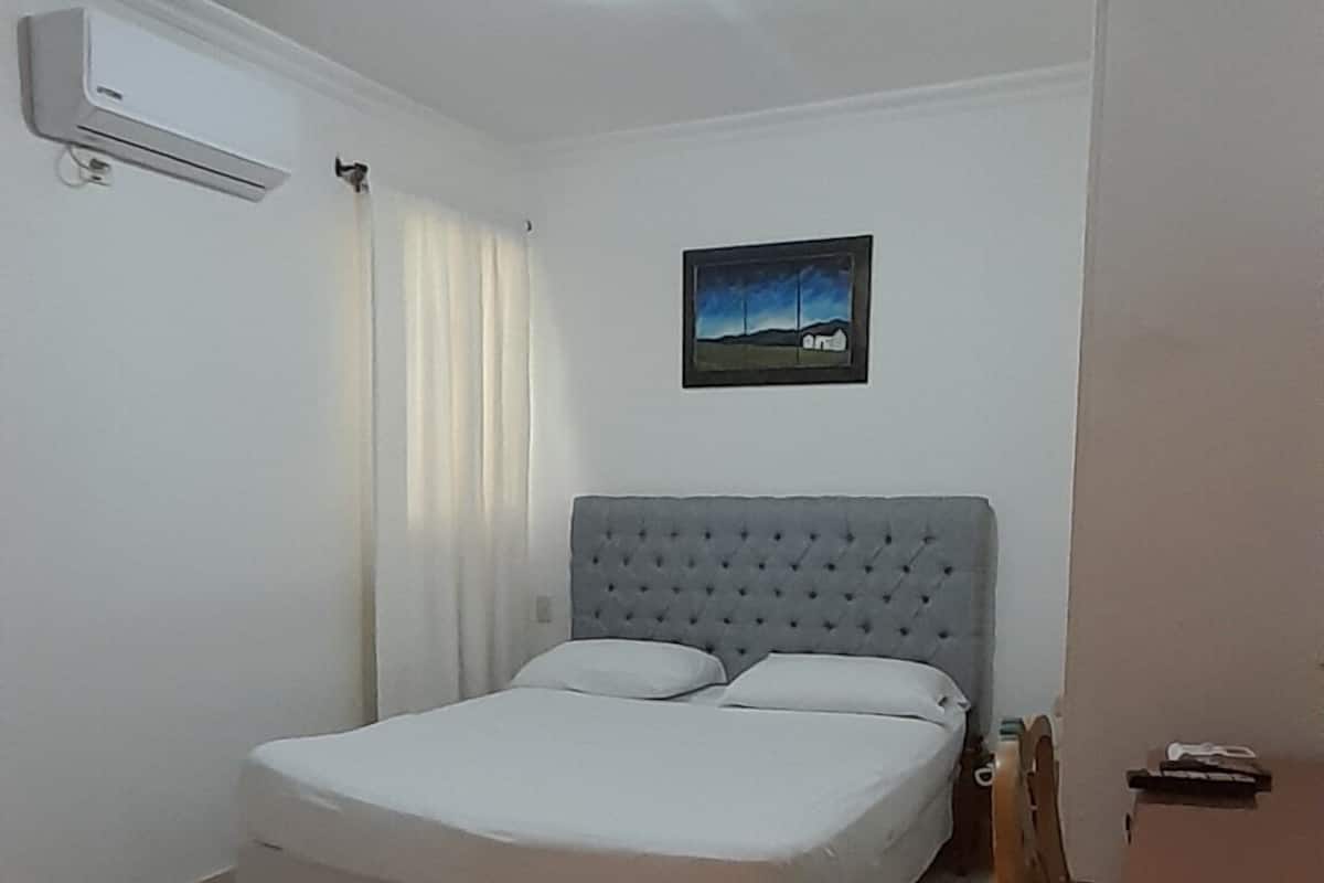 A bedroom with a bed, a desk, a chair, a picture on the wall, and an air conditioning unit.