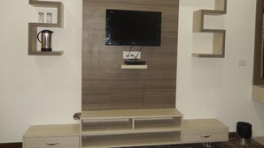 A modern living room with a flat-screen TV mounted on a wooden wall, a coffee maker on a shelf, and a minimalist entertainment unit. - Orchid Garden (New Delhi)
