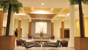 A spacious lobby with a modern design, featuring a central seating area with sofas and armchairs, flanked by tall palm trees in large planters. - The Grand Bhagwati Seasons Rajkot (Rajkot)