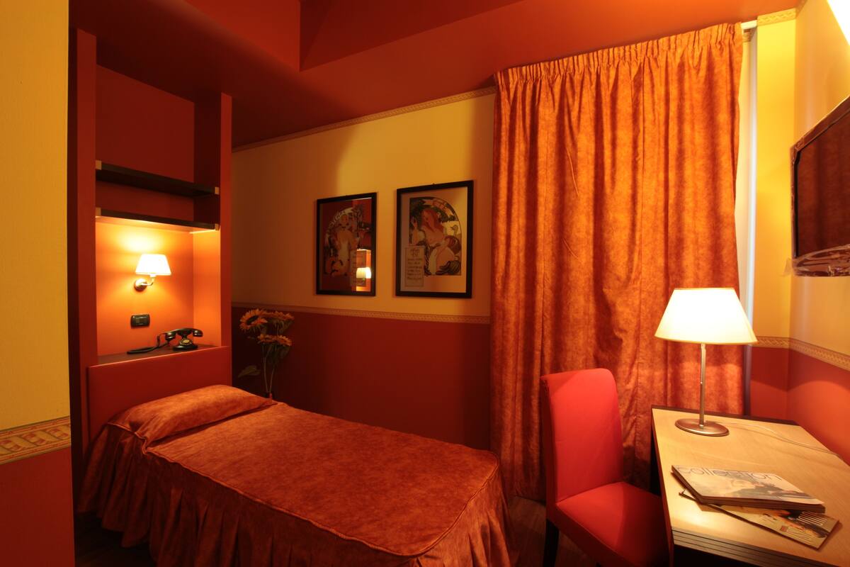 A hotel room with a bed, a desk with a lamp, a chair, and two framed pictures on the wall.