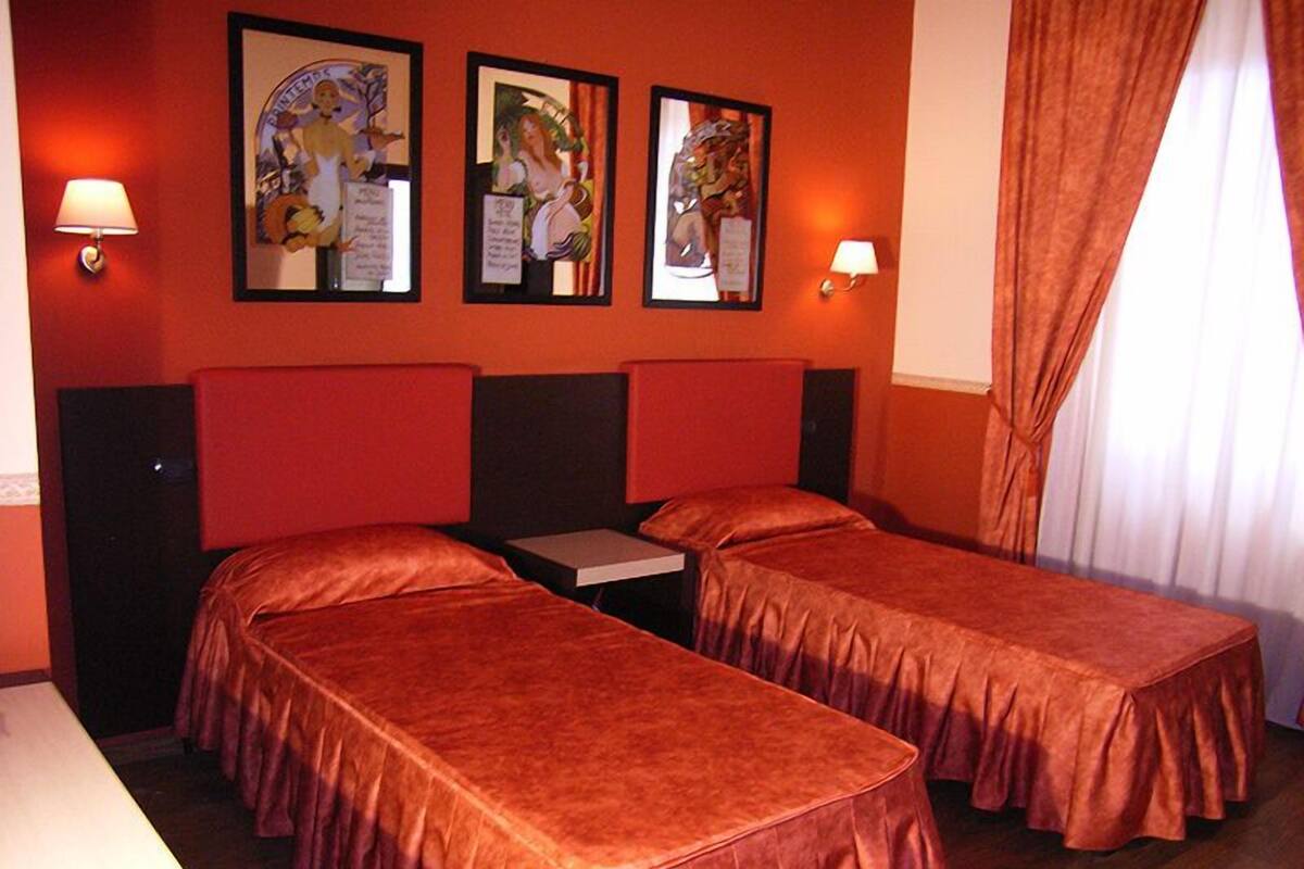 A hotel room with two single beds, a small table, and two wall-mounted lamps.