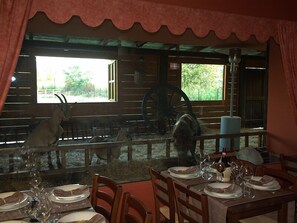 Un espace repas doté d’un décor intérieur rustique en bois, d’une grande fenêtre donnant sur un écurie où se trouvent des chevaux, et d’une table mise pour le repas.