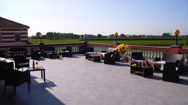 Un patio al aire libre con mesas y sillas, un jarrón decorativo con flores, y vistas a un campo verde y edificios a lo lejos.