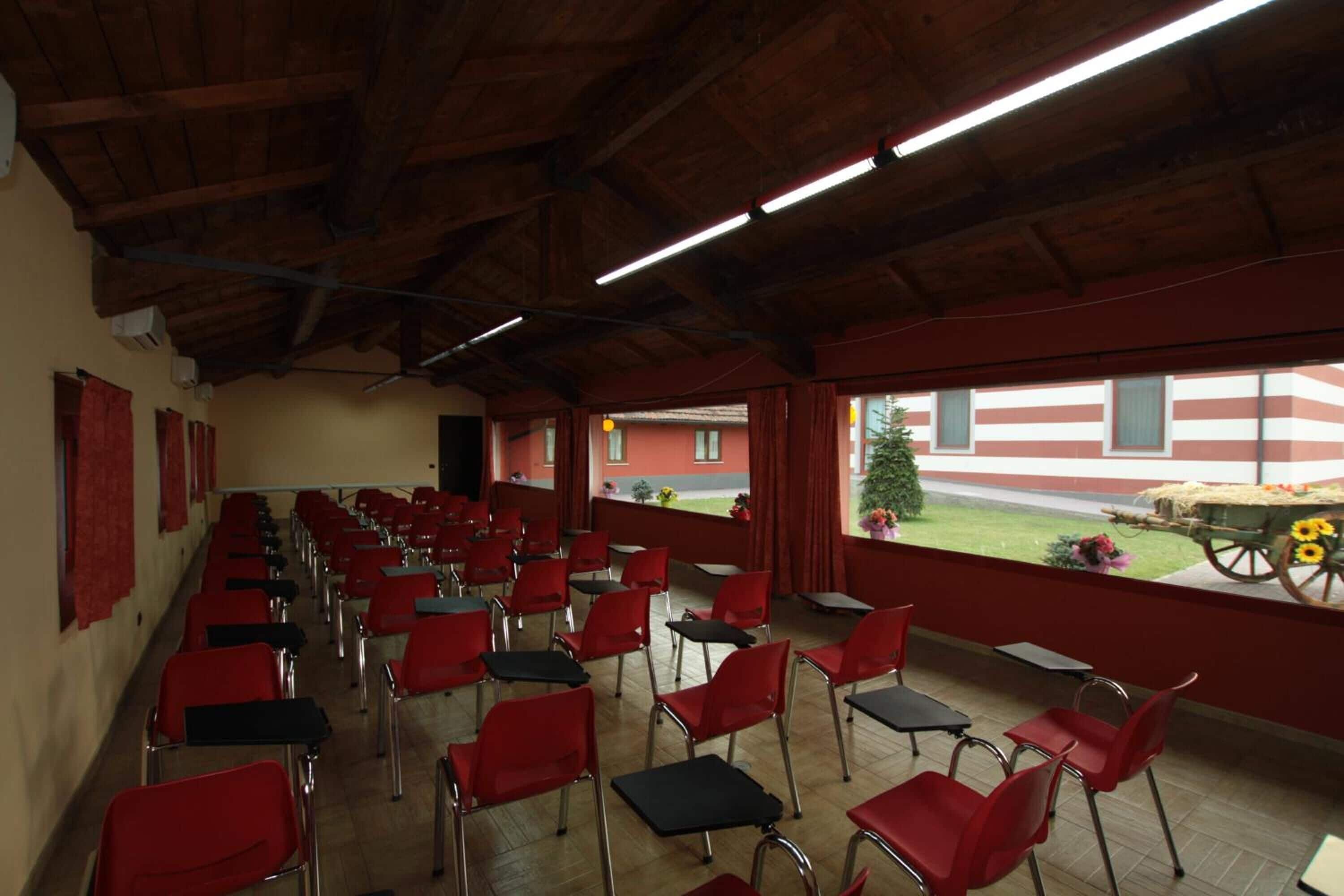 Un comedor con sillas y mesas rojas, vigas de madera en el techo y vistas a un edificio de fachada a rayas rojas y blancas.
