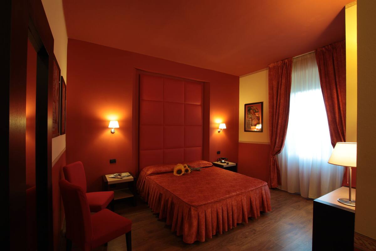 A hotel room with a large bed, two bedside tables, a chair, a window with curtains, and wall-mounted lamps.