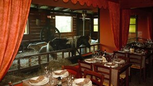 Un comedor con mesas dispuestas para una comida, vista de un establo con caballos, y un interior rústico con vigas de madera y cortinas rojas.