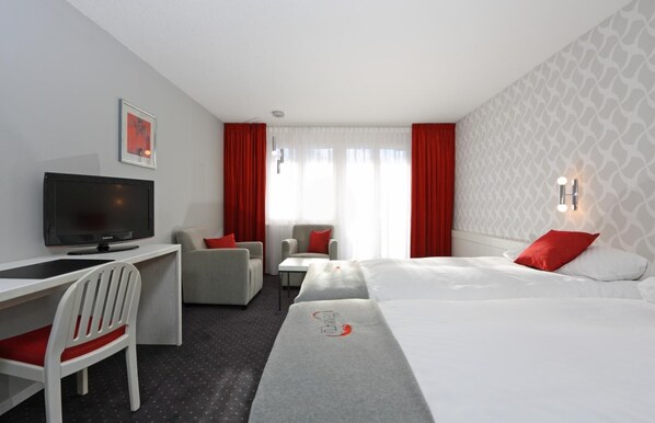 Habitación de hotel con cama, escritorio con televisor, silla, mesita y cortinas rojas.