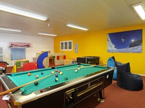 Una mesa de billar con bolas, una mesa de futbolín y un sillón de peluche en una habitación con paredes amarillas y una pintura de un paisaje montañoso.